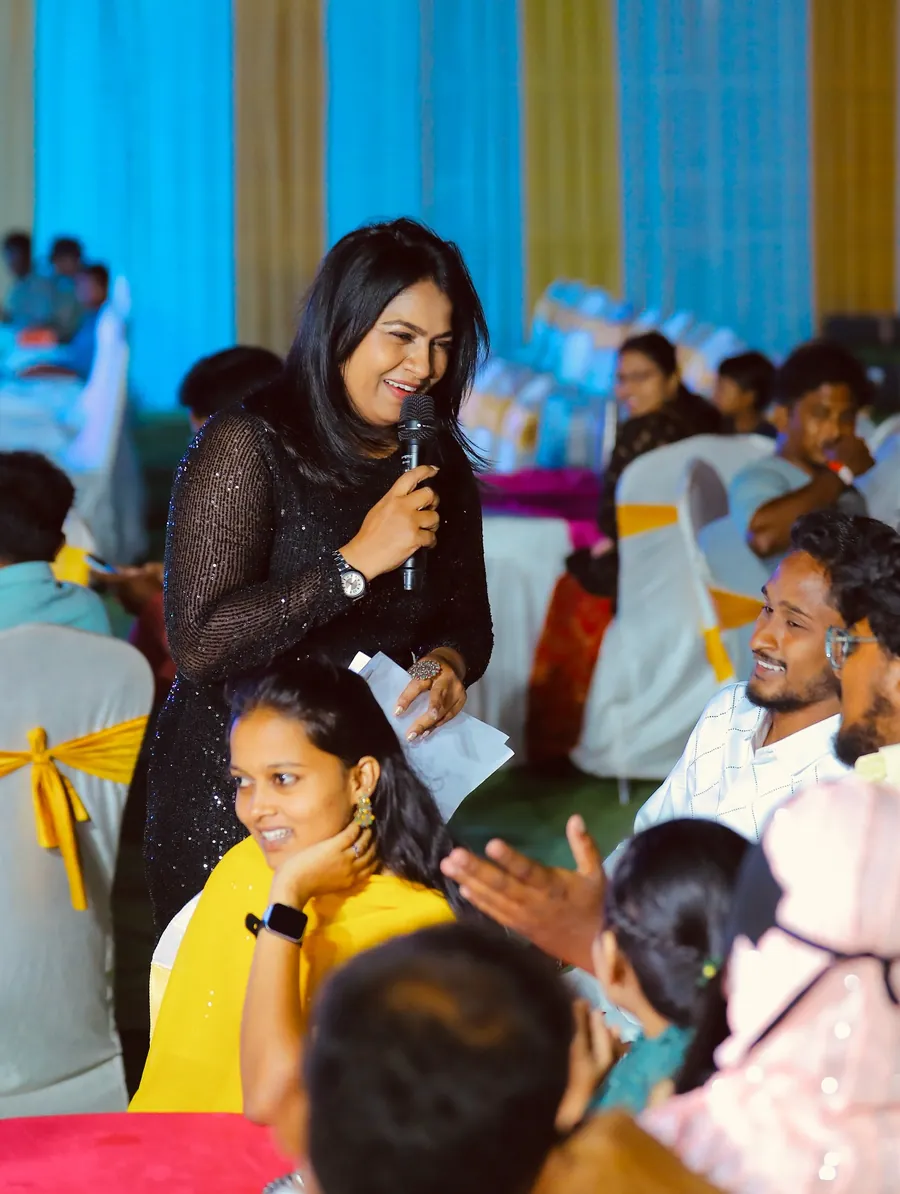 Reena with a microphone among the audience at a banquet, guests laughing along