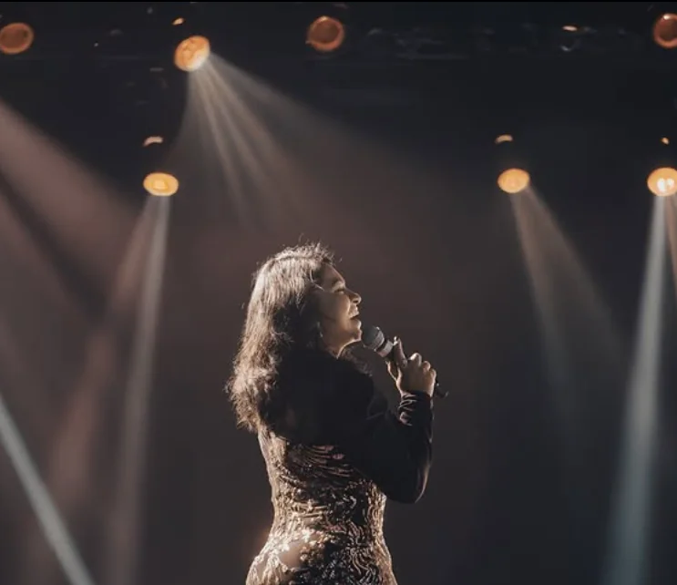 Reena singing into a microphone under warm stage spotlights